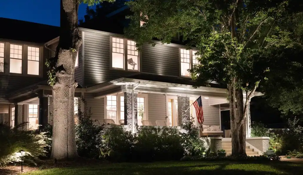 Outdoor Lighting Company in Waco TX Lumos Outdoor Lighting Suburban home at twilight with warm lighting highlighting the facade and surrounding trees. An American flag hangs on the porch, accentuating the home's welcoming feel.