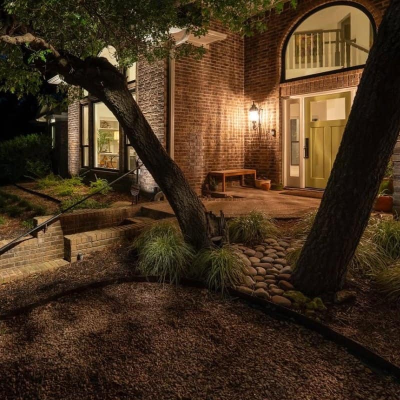 Outdoor Lighting Design near me 09 Exterior of a brick home at night, showcasing outdoor lighting design. A warm glow illuminates the front door and a bench, highlighting the landscaping and architectural details.