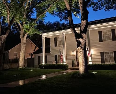 Outdoor Lighting Design near me 11 Elegant white home at dusk showcases professional outdoor lighting design, highlighting architectural details and mature trees. American flag displayed on the porch.