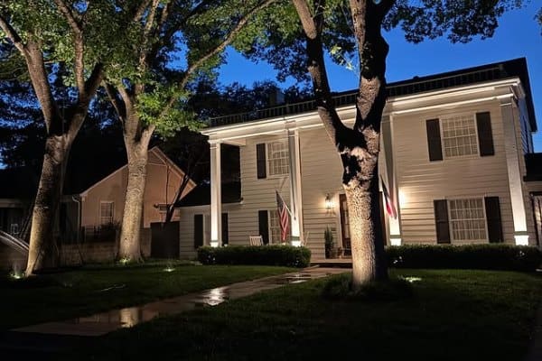 Outdoor Lighting Design near me 11 Elegant white home at dusk showcases professional outdoor lighting design, highlighting architectural details and mature trees. American flag displayed on the porch.
