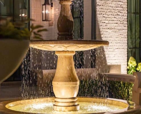 The Desco Outdoor Lighting Project Illuminated tiered stone fountain provides a focal point for outdoor lighting design, with water cascading into a circular basin, set against a brick facade and dark windows.