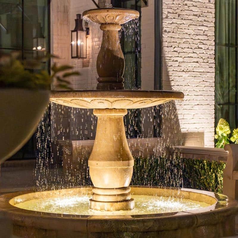 The Desco Outdoor Lighting Project Illuminated tiered stone fountain provides a focal point for outdoor lighting design, with water cascading into a circular basin, set against a brick facade and dark windows.