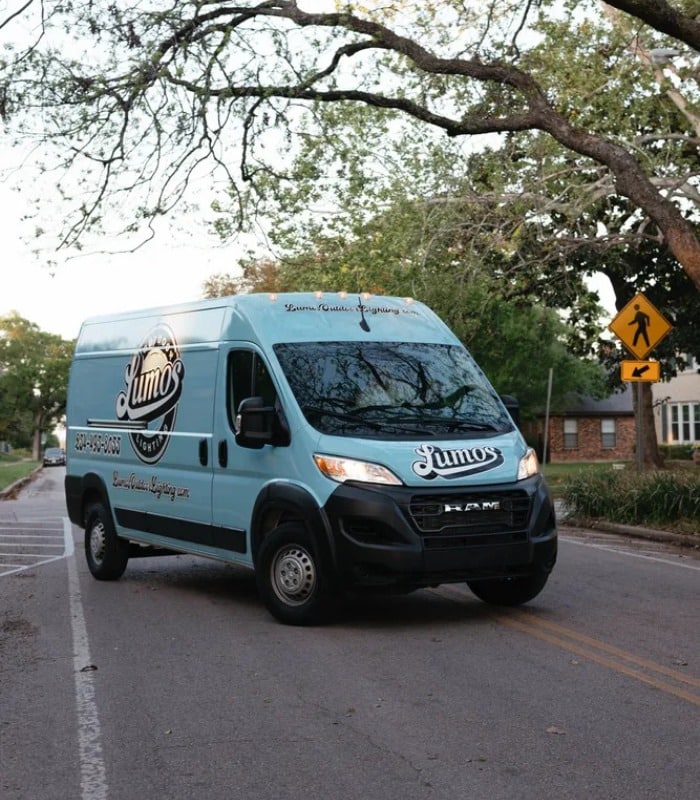 Outdoor Lighting Maintenance near me Waco TX 04 A light blue Lumos Lighting RAM van drives down a tree-lined street, promoting outdoor lighting maintenance services. The van prominently displays the company logo and contact information.