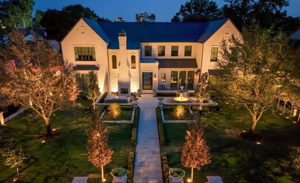 Outdoor Lighting Waco TX About Elegant white brick home illuminated by Lumos Lighting at dusk. The manicured lawn features a symmetrical garden path, mature trees, and a central fountain, all beautifully lit.