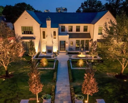 Elegant white brick home illuminated by Lumos Lighting at dusk. The manicured lawn features a symmetrical garden path, mature trees, and a central fountain, all beautifully lit.
