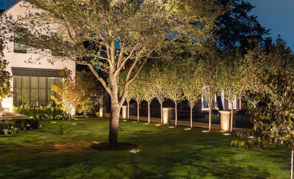 The Desco Outdoor Lighting Project Elegant landscape lighting installation illuminates a lush green lawn, highlighting trees, shrubs, and a brick fence. The soft, warm light creates a welcoming and sophisticated ambiance around the property.