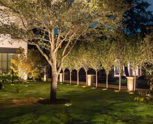 The Desco Outdoor Lighting Project Elegant landscape lighting installation illuminates a lush green lawn, highlighting trees, shrubs, and a brick fence. The soft, warm light creates a welcoming and sophisticated ambiance around the property.