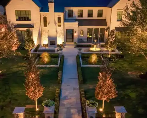 The Desco Outdoor Lighting Project Elegant home exterior featuring sophisticated outdoor lighting illuminating a manicured garden, stone pathway, and fountain.