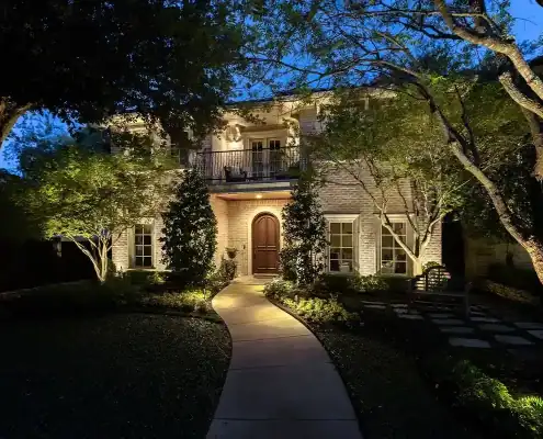 The Bunting Project Two-story brick house at dusk with warm exterior lights, curved path leading to an arched wooden front door, trees and landscaped yard.