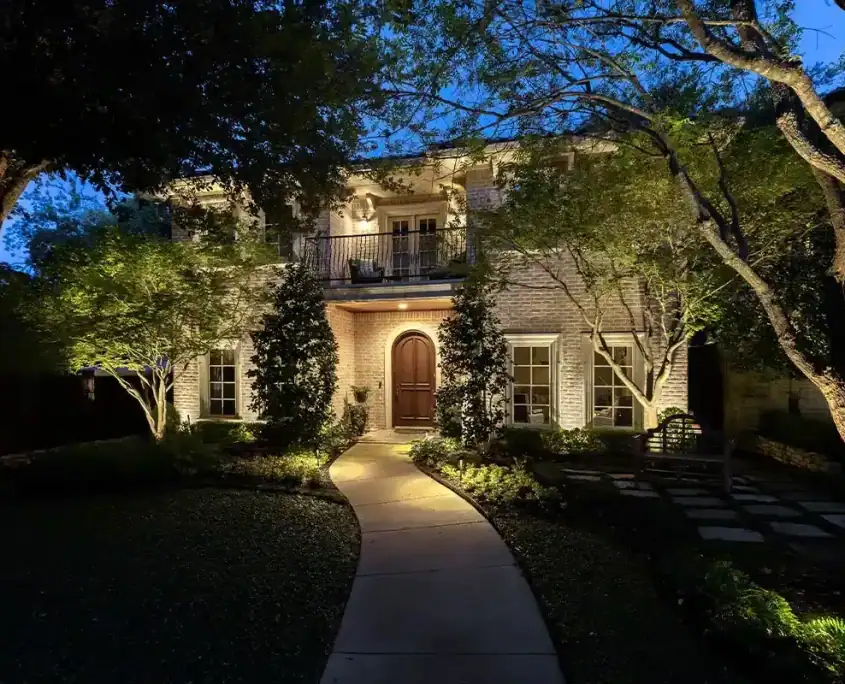 The Bunting Project Two-story brick house at dusk with warm exterior lights, curved path leading to an arched wooden front door, trees and landscaped yard.