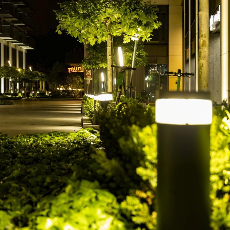Commercial-Landscape-Lighting-near-me-15 Nighttime commercial landscape lighting illuminates a pedestrian walkway. Modern light fixtures glow amidst lush greenery, enhancing the urban environment. A "muni" sign is visible in the background.