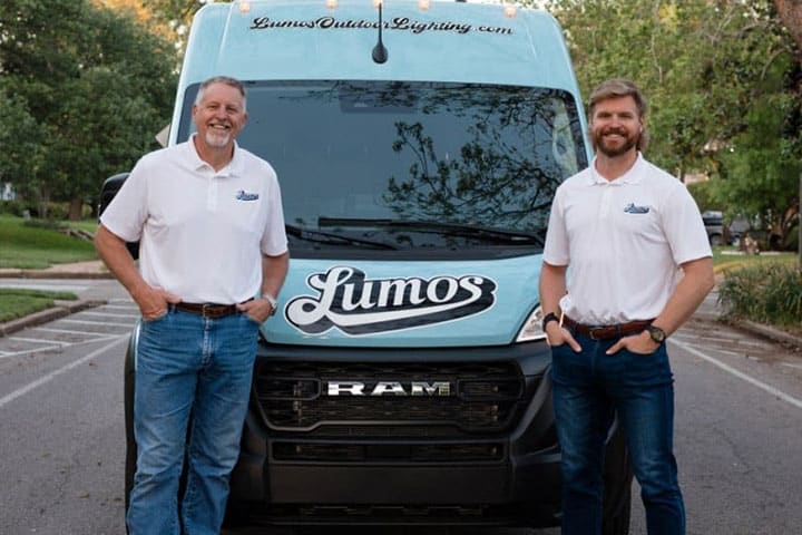 Outdoor-Lighting-Maintenance-near-me-Waco-TX-11 Two men stand proudly in front of a Lumos Outdoor Lighting van, promoting outdoor lighting maintenance services. The light blue van features the company logo and website.