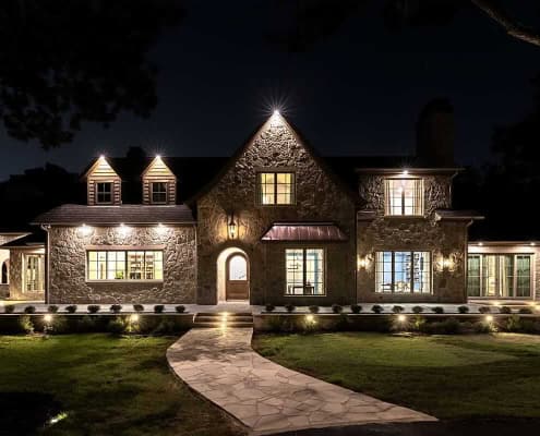 The Simondale Fort Worth Outdoor Lighting Project Large stone house at night, exterior lights and warm windows, curved stone walkway leading to arched front door and lawn.