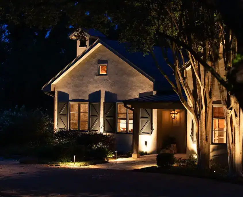 The Bear Waco Texas Outdoor Lighting Project Nighttime exterior of a stucco house with warm interior lights, closed shutters, a lit porch lantern, glowing pathway lights and nearby trees.