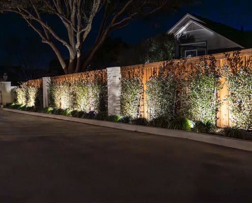 The Clover Ridge Outdoor Lighting Project Night view of a wooden fence along Clover Ridge, illuminated by landscape lighting. Green foliage grows along the fence, adding texture and depth to the scene.