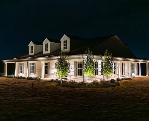The Clover Ridge Outdoor Lighting Project Clover Ridge at night: A single-story white house with dormers is illuminated against a dark sky, surrounded by a lawn and small trees.