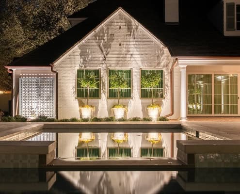 The Clover Ridge Outdoor Lighting Project Elegant home at Clover Ridge with a serene pool reflecting the illuminated white brick facade, windows, and potted trees. Soft lighting enhances the tranquil ambiance.