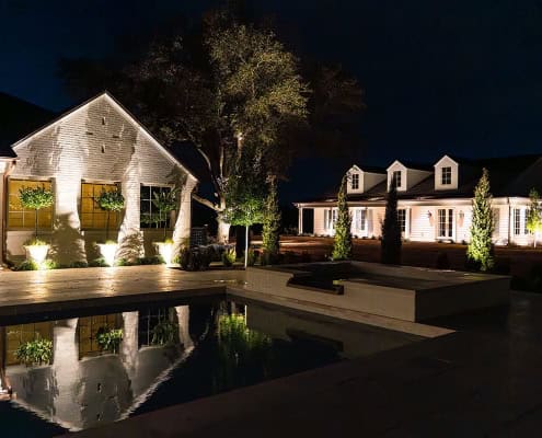 The Clover Ridge Outdoor Lighting Project Night view of Clover Ridge estate featuring a pool reflecting the illuminated white farmhouse architecture. Outdoor lighting accentuates the manicured landscaping and modern design.