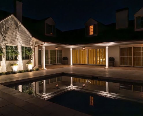 The Clover Ridge Outdoor Lighting Project Night view of Clover Ridge's serene pool reflecting the illuminated architecture. The white brick home with dark roof and dormers creates a calming, elegant ambiance.
