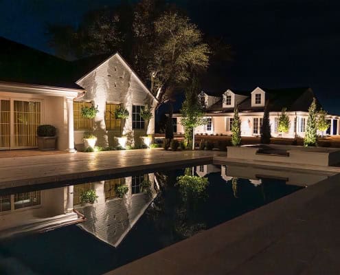 The Clover Ridge Outdoor Lighting Project Clover Ridge estate at night, reflected in a serene pool. White farmhouse architecture is illuminated by landscape lighting, showcasing the property's elegant design and tranquil ambiance.
