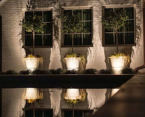 The Clover Ridge Outdoor Lighting Project Exterior of Clover Ridge home at night. Three potted topiary trees are illuminated against a white brick wall, reflected in a pool. Modern and elegant outdoor design.