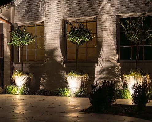The Clover Ridge Outdoor Lighting Project Exterior of the Clover Ridge house at night, showcasing landscape lighting illuminating potted trees and the brick facade. Warm light glows through the windows.