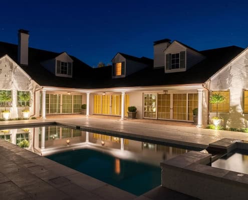 The Clover Ridge Outdoor Lighting Project Luxury home at night with a pool reflecting the house lights under a starry sky. The white painted brick exterior of Clover Ridge glows warmly around the courtyard.