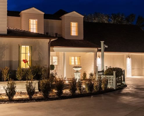 The Clover Ridge Outdoor Lighting Project Exterior twilight view of a white brick home with a dark roof. Warm light shines from the windows, illuminating manicured landscaping and a circular driveway at Clover Ridge.
