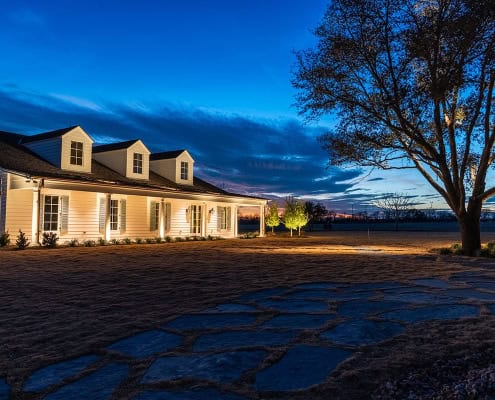 The Clover Ridge Outdoor Lighting Project Clover Ridge residence at dusk: A white farmhouse with dormers glows under soft lighting against a twilight sky, framed by a large tree and a stone pathway.