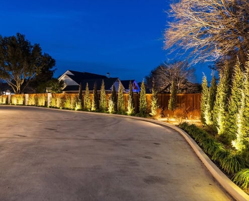 The Clover Ridge Outdoor Lighting Project Night view of Clover Ridge landscaping, featuring illuminated trees and a curved driveway. Warm lights highlight the fence and greenery, creating an inviting, upscale ambiance.