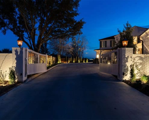 The Clover Ridge Outdoor Lighting Project An illuminated driveway leads through white brick gates into the Clover Ridge estate at dusk. The long driveway is lined with trees and landscaping, creating an inviting entrance.