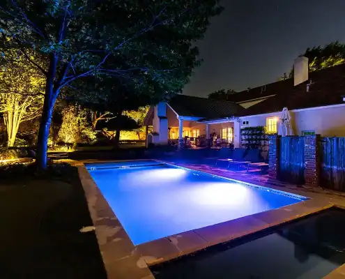 The Colonial Fort Worth Outdoor Lighting Project Night backyard with a glowing blue rectangular pool, patio loungers, illuminated trees and a house with warm-lit windows.