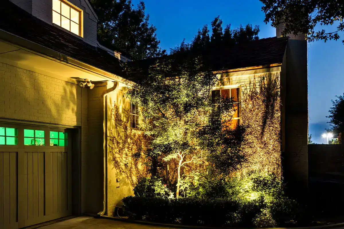 The Colonial Fort Worth Outdoor Lighting Project Night view of a house exterior with a lit tree and shrubs casting shadows on a brick wall, illuminated windows and garage door with green panes.