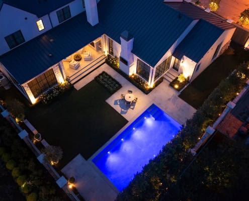 The Desco Outdoor Lighting Project Aerial view of a modern luxury home at night. Features include a rectangular pool with blue lighting, a patio dining set, manicured lawns, and exterior lighting highlighting the architecture.