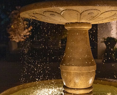 The Desco Outdoor Lighting Project Illuminated stone fountain at night, water droplets suspended in air. A serene scene, perfect for outdoor lighting projects, showcasing the gentle glow and ambiance.