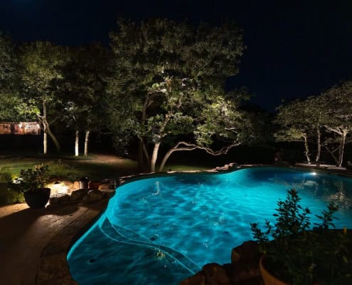 The Poolside Serenity—Moonlit Canopy Design Nighttime view of a turquoise-lit pool surrounded by lush trees illuminated by landscape lighting. The scene evokes a sense of poolside serenity under a moonlit canopy design.