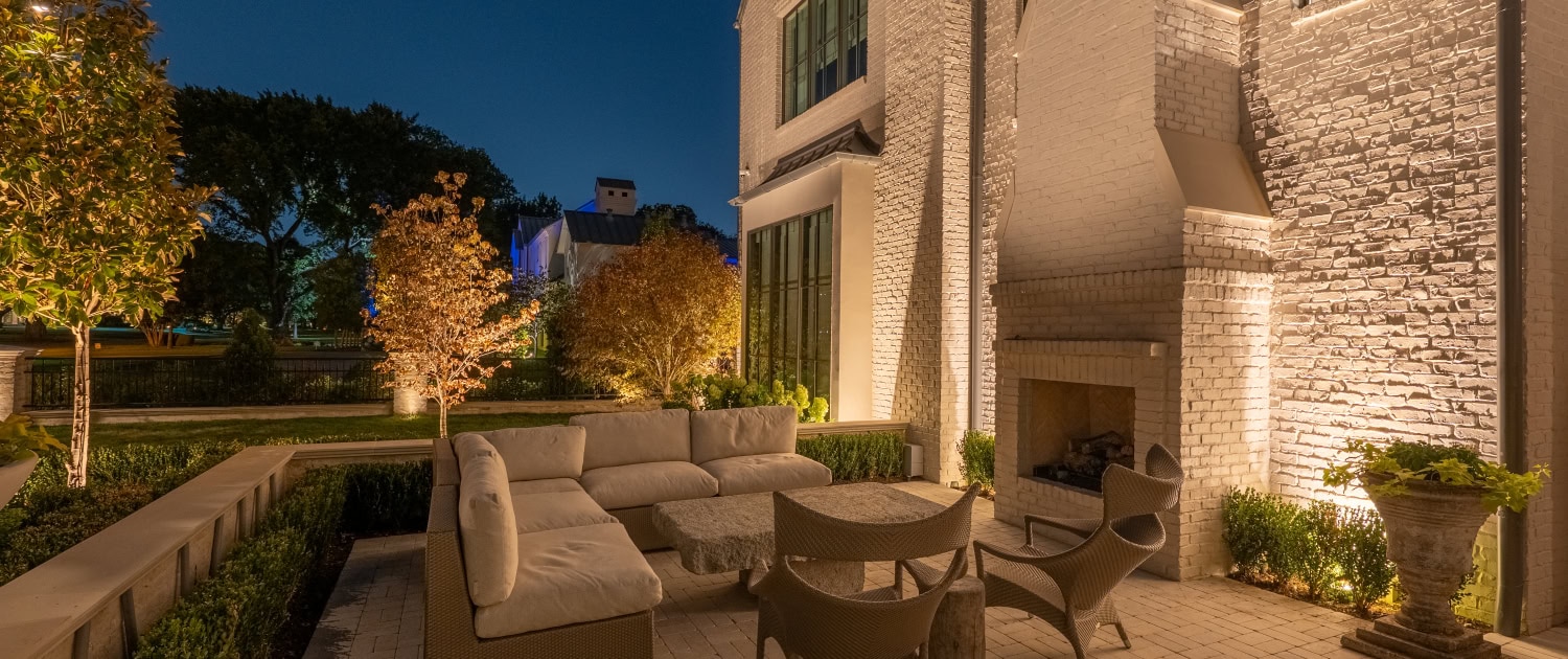 Lumos Outdoor Lighting Patio Scene Elegant outdoor patio illuminated by Lumos outdoor lighting, featuring a cozy seating area with a fireplace, surrounded by lush greenery and trees at dusk.