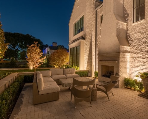Lumos Outdoor Lighting Patio Scene Elegant outdoor patio illuminated by Lumos outdoor lighting, featuring a cozy seating area with a fireplace, surrounded by lush greenery and trees at dusk.