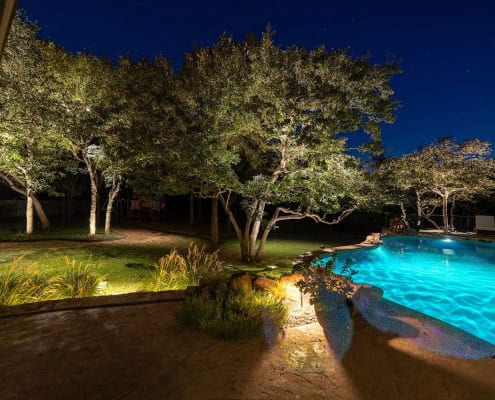 The Poolside Serenity—Moonlit Canopy Design Night view of a backyard pool illuminated with turquoise lighting, reflecting the moonlit canopy design of trees spotlighted around the yard. Patio and lush landscaping surround the pool.