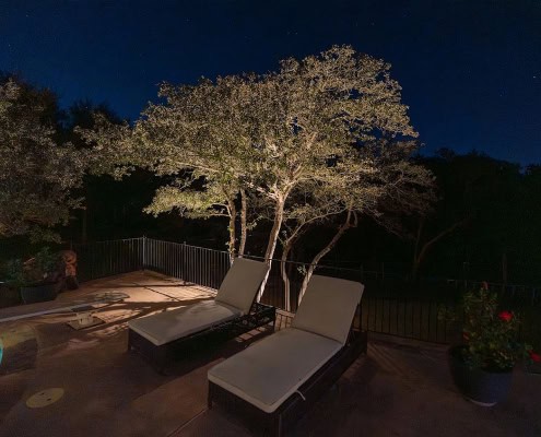 The Poolside Serenity—Moonlit Canopy Design Two lounge chairs sit poolside under a tree illuminated by outdoor lighting at night, creating a serene moonlit canopy design. The pool glows blue, and stars are visible in the dark sky.