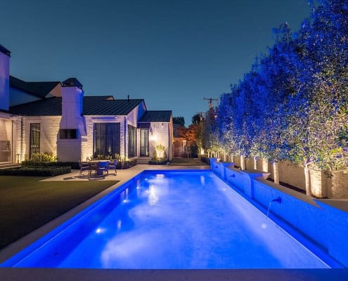 The Desco Outdoor Lighting Project Luxury backyard pool at twilight, reflecting blue light. Modern white brick home, manicured lawn, and trees with upward lighting create a serene "Poolside Serenity" design.