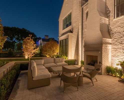 The Desco Outdoor Lighting Project Outdoor patio at dusk featuring a cream-colored sectional sofa, wicker chairs, and a stone-topped table, illuminated by landscape lighting. A brick fireplace and manicured hedges add to the elegant ambiance of The Desco Project design.