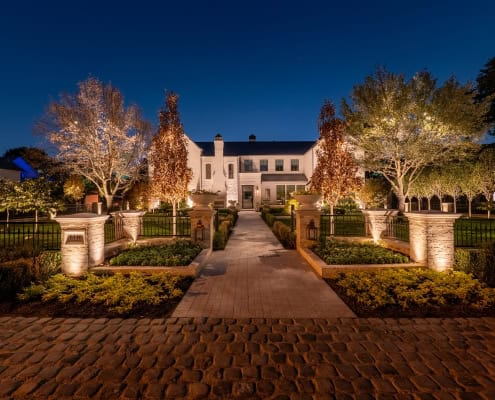 The Desco Outdoor Lighting Project Elegant estate illuminated at night. Stone walkway leads to a grand white house with manicured landscaping, including trees adorned with lights. Part of The Desco Project, showcasing sophisticated outdoor lighting design.