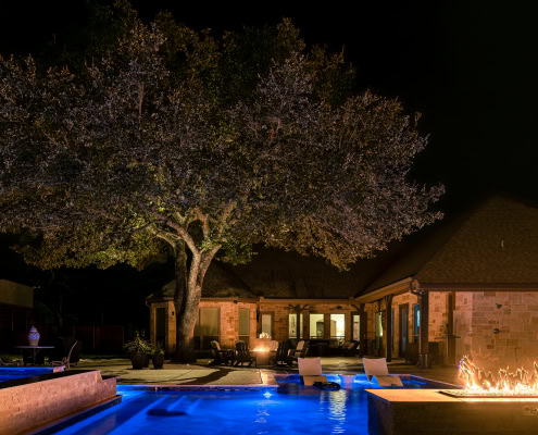 Nighttime view of a beautifully lit outdoor pool area with landscape lighting, fire feature, and cozy seating under a large tree.