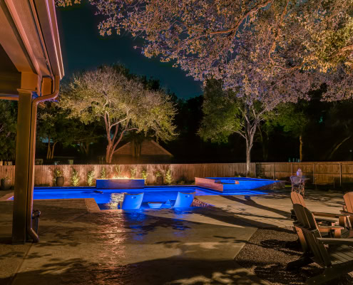 Night view of a backyard with illuminated pool, trees, and outdoor seating, showcasing Lumos Outdoor Lighting's landscape lighting solutions.