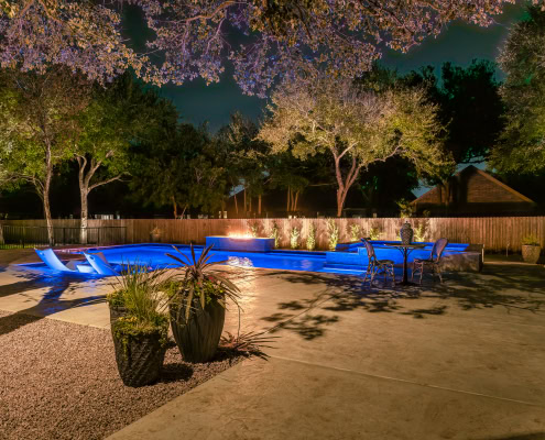 Night view of a backyard with illuminated pool, outdoor seating, and landscape lighting by Lumos Outdoor Lighting.
