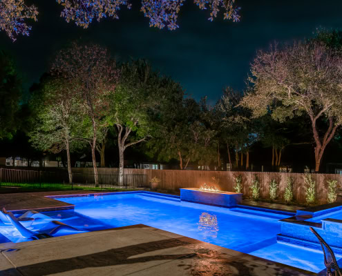 Night view of a backyard pool illuminated by Lumos Outdoor Lighting, showcasing vibrant blue water and landscape lighting among trees.