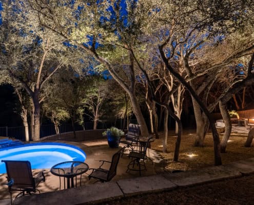 outdoor-lighting-pool-trees Outdoor lighting softly illuminating trees around a patio with a fire pit on the right side and an illuminated pool with patio chairs surrounding it on the left.