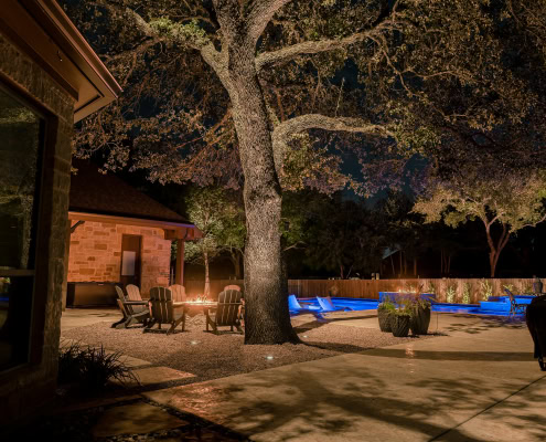 Nighttime outdoor lighting illuminating a backyard with a pool, trees, and patio furniture, showcasing professional landscape lighting design by Lumos Outdoor Lighting.