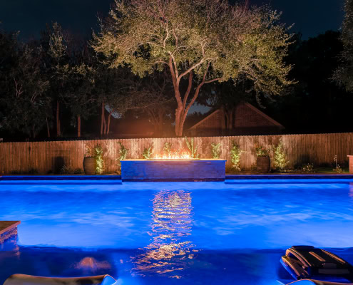 Night view of a backyard pool illuminated by Lumos outdoor lighting, showcasing vibrant blue water and landscape lighting highlighting trees and garden features.